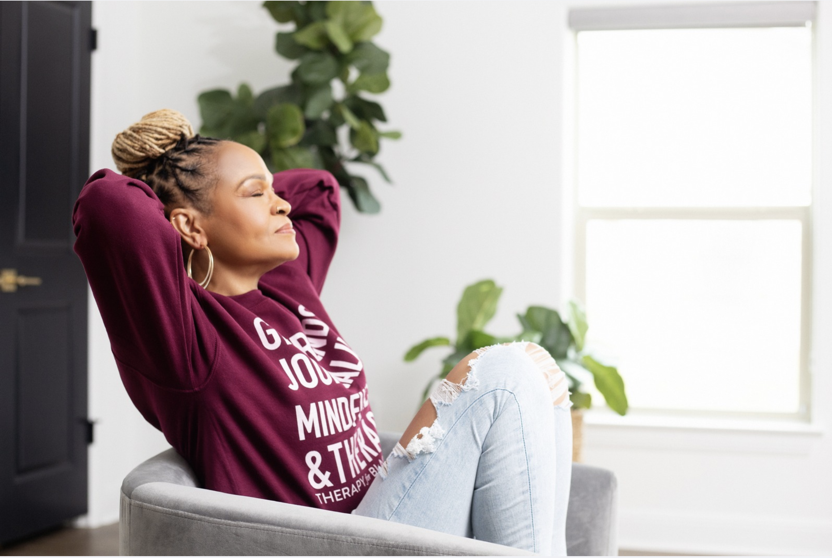 A woman with braided hair in a bun sits relaxed in a grey chair, wearing a burgundy sweatshirt and ripped jeans, with her eyes closed and hands behind her head, symbolizing a moment of peace and reflection in the journey of rewriting her internal script